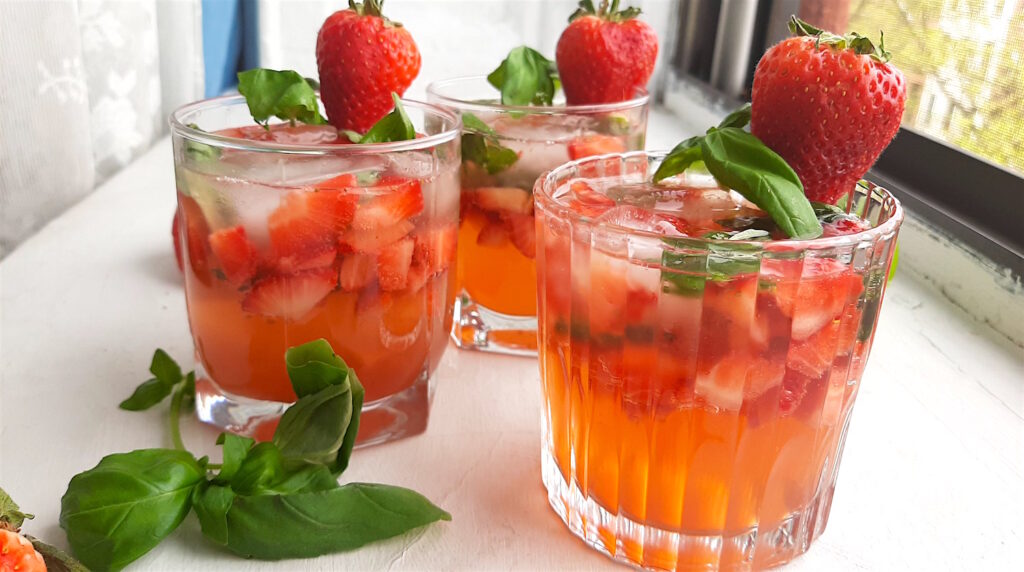 Balsamic Strawberry Basil cocktail, three strawberry basil cocktails in tumblers on a white background. Garnished with fresh basil and strawberries. Surrounded by limes and basil. Front Shot. Sugar with Spice Blog.
