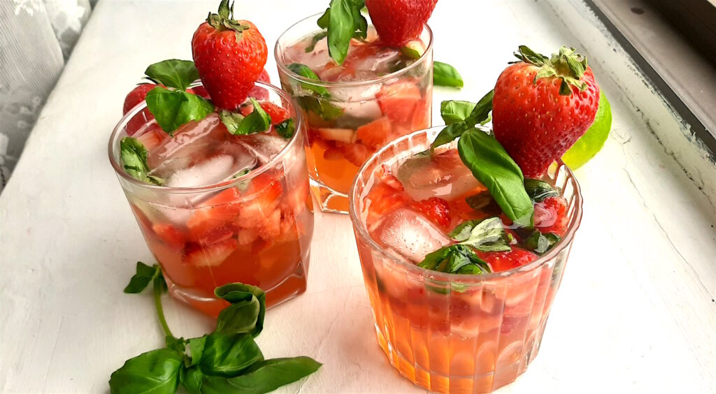 Balsamic Strawberry Basil cocktail, three strawberry basil cocktails in tumblers on a white background. Garnished with fresh basil and strawberries. Surrounded by limes and basil. Far overhead wide shot. Sugar with Spice Blog.