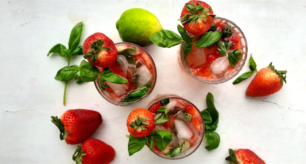 Balsamic Strawberry Basil cocktail, three strawberry basil cocktails in tumblers on a white background. Garnished with fresh basil and strawberries. Surrounded by limes and basil. Overhead wide shot. Sugar with Spice Blog.