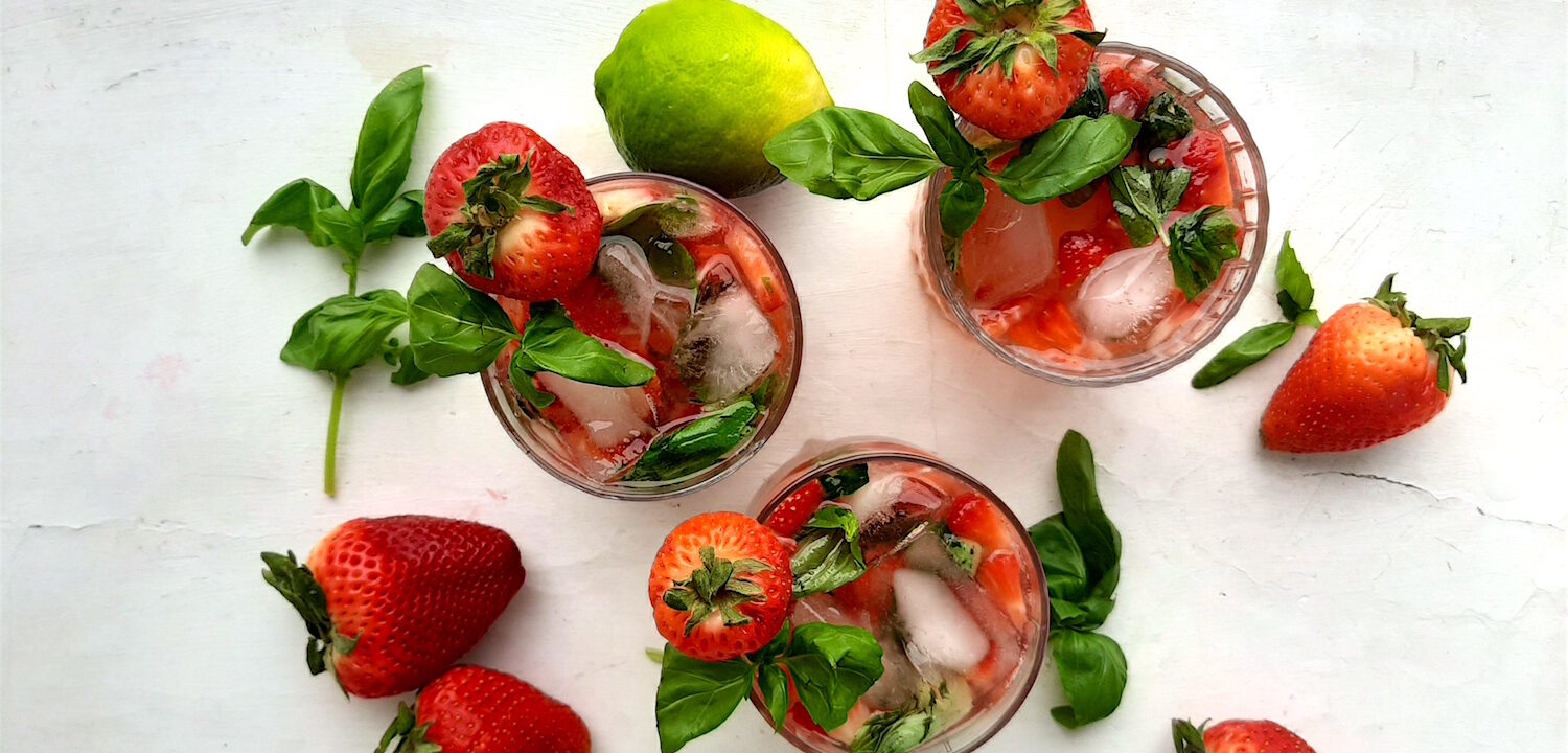 Balsamic Strawberry Basil cocktail, three strawberry basil cocktails in tumblers on a white background. Garnished with fresh basil and strawberries. Surrounded by limes and basil. Overhead wide shot. Sugar with Spice Blog.