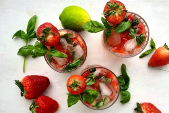 Balsamic Strawberry Basil cocktail, three strawberry basil cocktails in tumblers on a white background. Garnished with fresh basil and strawberries. Surrounded by limes and basil. Overhead wide shot. Sugar with Spice Blog.