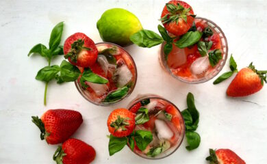 Balsamic Strawberry Basil cocktail, three strawberry basil cocktails in tumblers on a white background. Garnished with fresh basil and strawberries. Surrounded by limes and basil. Overhead wide shot. Sugar with Spice Blog.