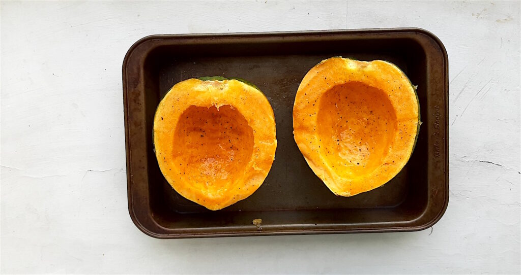 Acorn Squash ready to be roasted, sliced in half-lengthways in a metal baking dish on a white background.