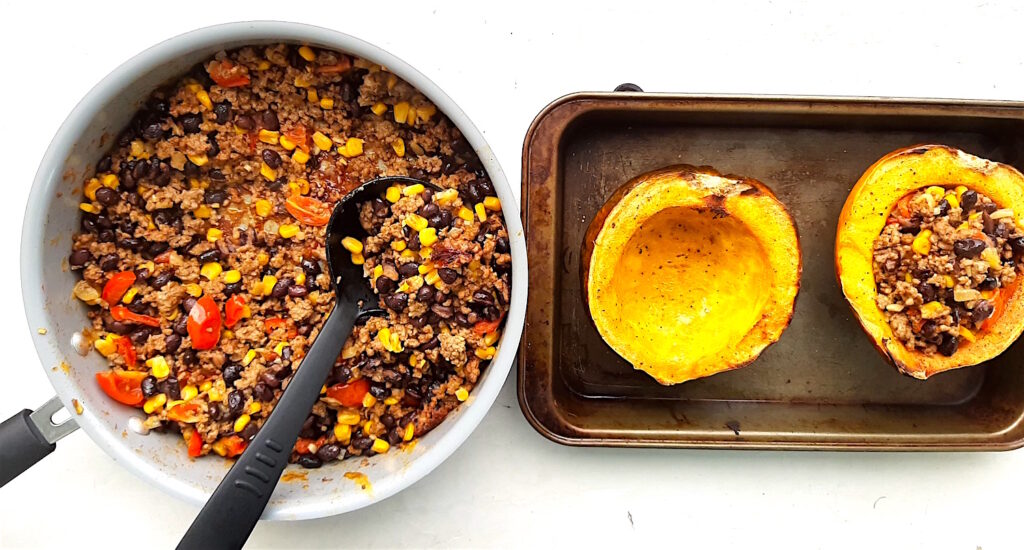 Mexican-style stuffed acorn squash being filled. Two halves of baked acorn squash in a baking dish. Pan of Mexican-style filling nearby with a black serving spoon. One squash half is filled. White background. Sugar with Spice Blog.