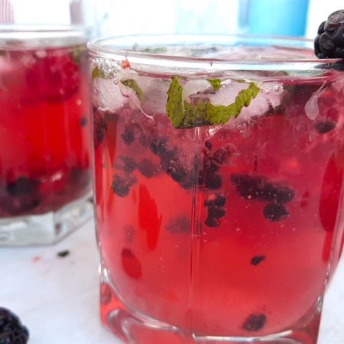 Three Mixed Berry Mojitos in tumbler glasses, garnished with blackberries, raspberries, and mint. On a white window sill surrounded by fresh blackberries, raspberries, cardamom pods. Sugar with Spice Blog.