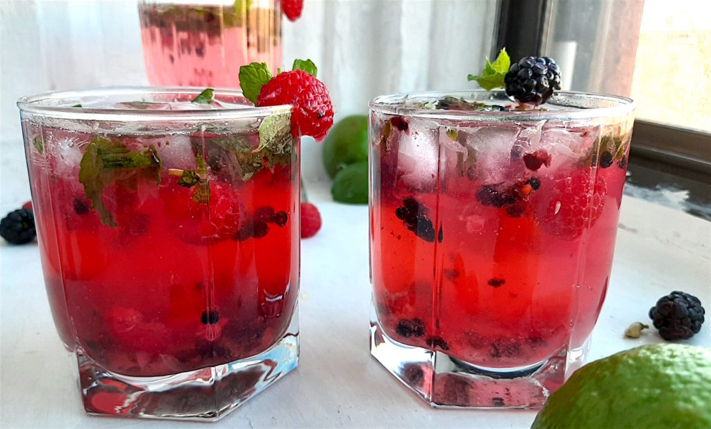 Three Mixed Berry Mojitos in tumbler glasses, garnished with blackberries, raspberries, and mint. On a white window sill surrounded by fresh blackberries, raspberries, cardamom pods. Sugar with Spice Blog.