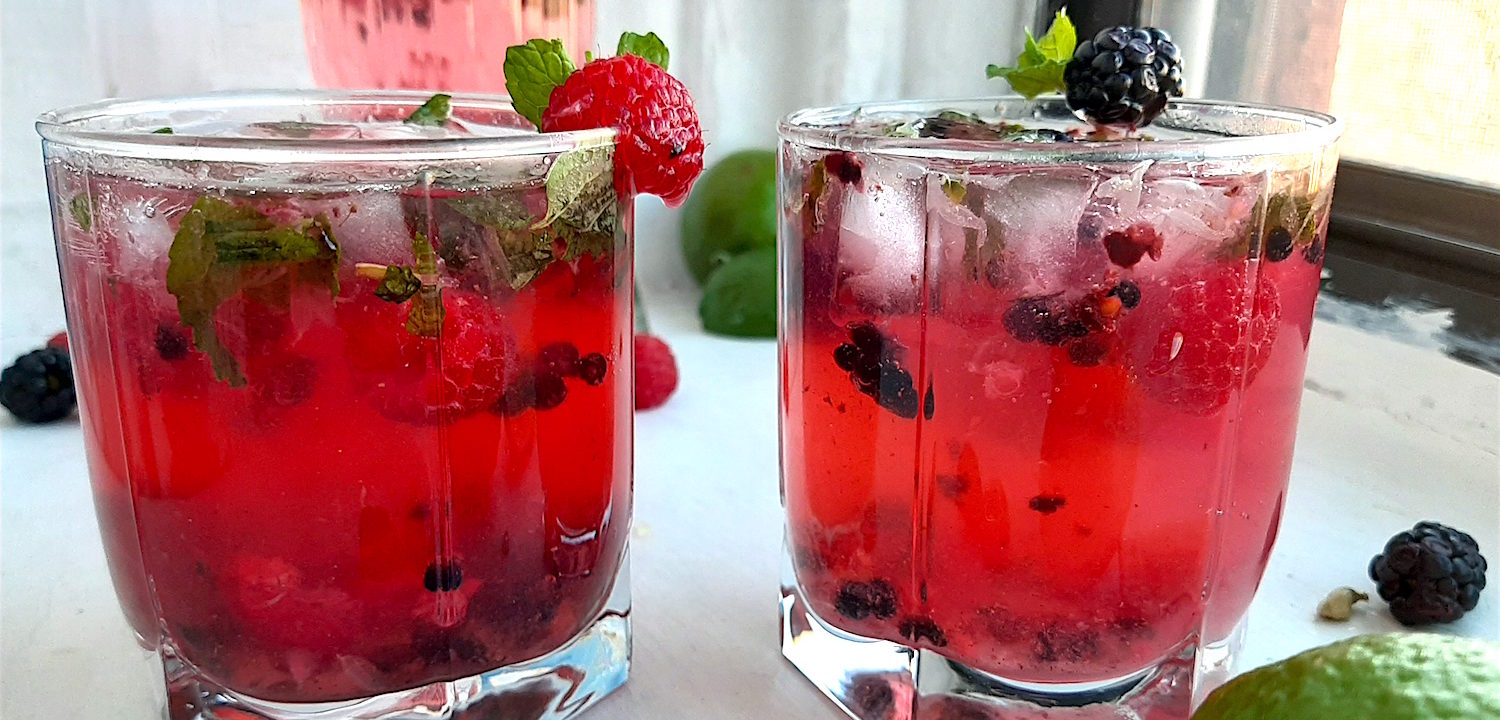 Three Mixed Berry Mojitos in tumbler glasses, garnished with blackberries, raspberries, and mint. On a white window sill surrounded by fresh blackberries, raspberries, cardamom pods. Sugar with Spice Blog.