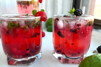 Three Mixed Berry Mojitos in tumbler glasses, garnished with blackberries, raspberries, and mint. On a white window sill surrounded by fresh blackberries, raspberries, cardamom pods. Sugar with Spice Blog.