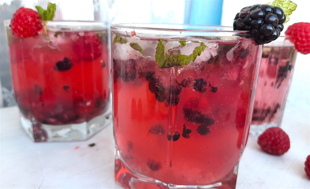 Three Mixed Berry Mojitos in tumbler glasses, garnished with blackberries, raspberries, and mint. On a white window sill surrounded by fresh blackberries, raspberries, cardamom pods. Sugar with Spice Blog.