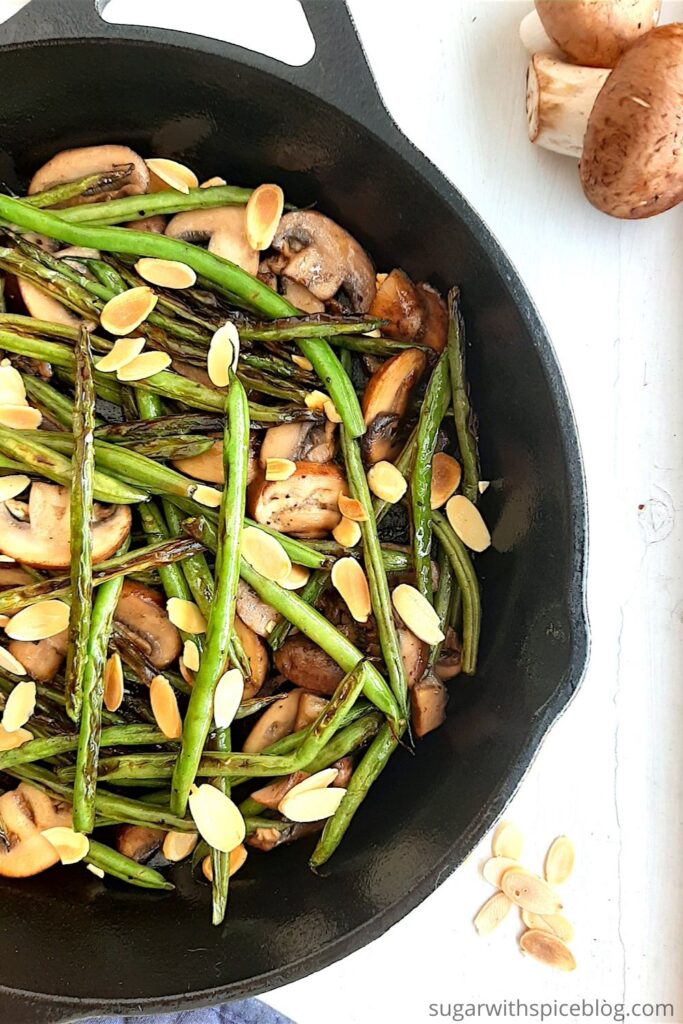 Almond and Mushroom Green Beans in a cast iron skillet surrounded by a blue oven mitt, fresh mushrooms, and sliced almonds. Wide overhead shot pinterest image. Sugar with Spice Blog.