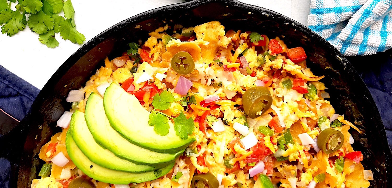Classic Tex-Mex Migas topped with avocado, pickled jalapeños, and sliced avocado in a cast iron skillet on a white background with blue oven mitts.. Overhead off kilter shot. Sugar with Spice Blog.