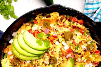 Classic Tex-Mex Migas topped with avocado, pickled jalapeños, and sliced avocado in a cast iron skillet on a white background with blue oven mitts.. Overhead off kilter shot. Sugar with Spice Blog.