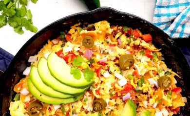 Classic Tex-Mex Migas topped with avocado, pickled jalapeños, and sliced avocado in a cast iron skillet on a white background with blue oven mitts.. Overhead off kilter shot. Sugar with Spice Blog.