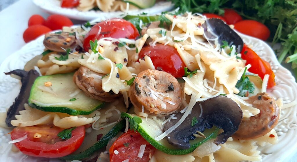 Smoked Sausage and Veggie Bow Tie Pasta Skillet with bow tie pasta, smoked sausage slices, mushrooms, zucchini slices, and halved cherry tomatoes on two white plates. Fresh parsley and cherry tomatoes to the side. White background, close up front shot. Sugar with Spice Blog.