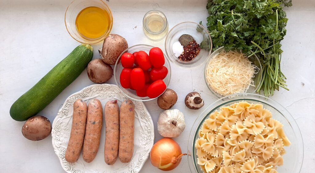 Smoked Sausage and Veggie Bow Tie Pasta Skillet ingredients. Smoked Chicken sausages on a white plate, cooked bow tie pasta in a pyrex dish, Whole zucchini, cherry tomatoes, garlic, white onion, and mushrooms with fresh parsley. Olive oil and parmesan cheese in small pyrex bowls, a small carafe of white wine all on a white background overhead shot. Sugar with Spice Blog.