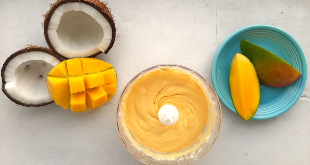 Mango Coconut Soft Serve Ice Cream mixed in a blender with fresh sliced mango and split coconut to one side. The other side has a blue plate with sliced mango pieces. All on a white background, overhead shot. Sugar with Spice Blog.