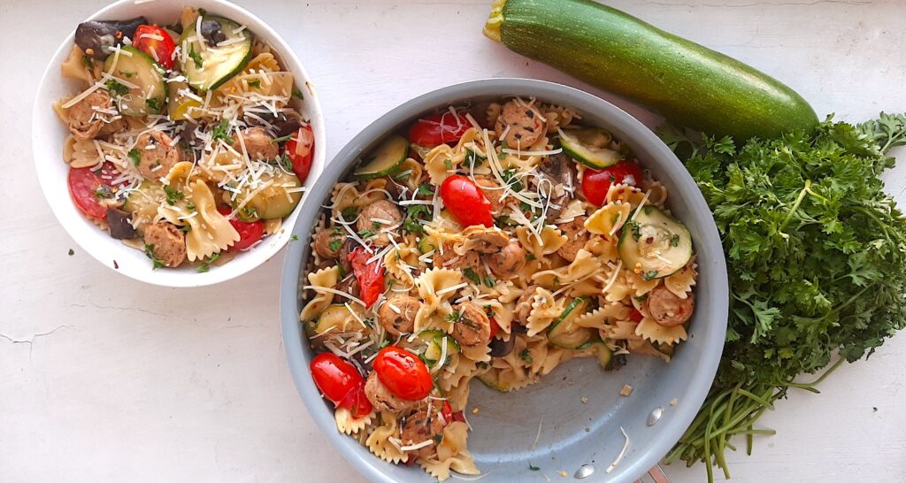 Smoked Sausage and Veggie Bow Tie Pasta Skillet with bow tie pasta, smoked sausage slices, mushrooms, zucchini slices, and halved cherry tomatoes in a medium sauté pan. A white bowl with more pasta, sausage, and veggies to the side. Fresh Parsley and a whole zucchini to the other side. White background overhead shot. Sugar with Spice Blog.