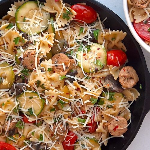 Smoked Sausage and Veggie Bow Tie Pasta Skillet with bow tie pasta, smoked sausage slices, mushrooms, zucchini slices, and halved cherry tomatoes in a medium cast iron skillet. A white bowl with more pasta, sausage, and veggies to the side. Fresh Parsley to the other side. White background. Sugar with Spice Blog.