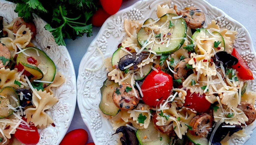 Smoked Sausage and Veggie Bow Tie Pasta Skillet with bow tie pasta, smoked sausage slices, mushrooms, zucchini slices, and halved cherry tomatoes on two white plates. Fresh parsley and cherry tomatoes to the side. White background, overhead shot. Sugar with Spice Blog.