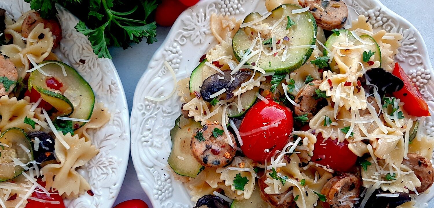 Smoked Sausage and Veggie Bow Tie Pasta Skillet with bow tie pasta, smoked sausage slices, mushrooms, zucchini slices, and halved cherry tomatoes on two white plates. Fresh parsley and cherry tomatoes to the side. White background, overhead shot. Sugar with Spice Blog.