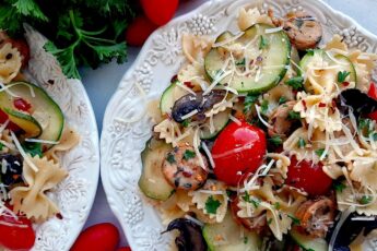 Smoked Sausage and Veggie Bow Tie Pasta Skillet with bow tie pasta, smoked sausage slices, mushrooms, zucchini slices, and halved cherry tomatoes on two white plates. Fresh parsley and cherry tomatoes to the side. White background, overhead shot. Sugar with Spice Blog.