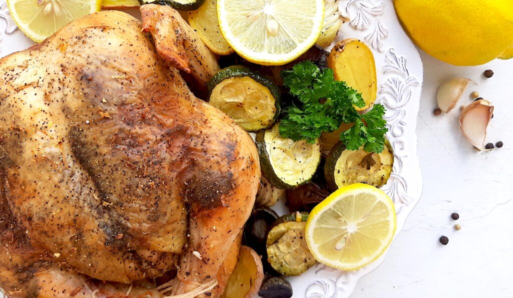 Lemon Pepper Cornish Hen on a white plate with assorted mixed veggies like potatoes, zucchini rounds, mushrooms, and onions on the side. Lemon rounds and parsley for garnish. Whole lemons, garlic cloves, and pepper kernels on the side, white background. Overhead shot. Sugar with Spice Blog.