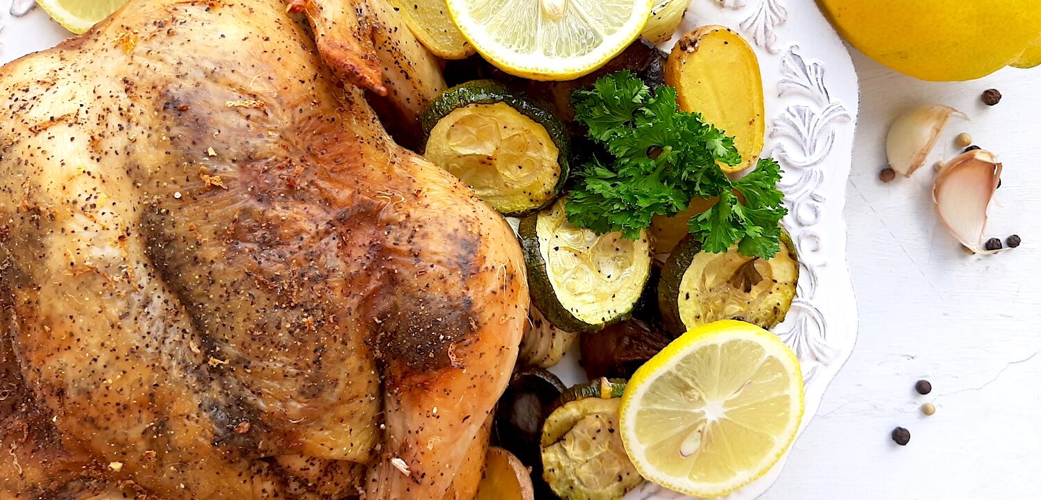 Lemon Pepper Cornish Hen on a white plate with assorted mixed veggies like potatoes, zucchini rounds, mushrooms, and onions on the side. Lemon rounds and parsley for garnish. Whole lemons, garlic cloves, and pepper kernels on the side, white background. Overhead shot. Sugar with Spice Blog.