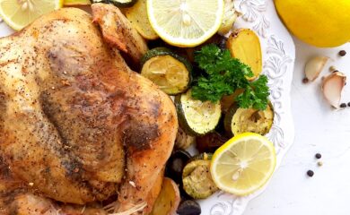 Lemon Pepper Cornish Hen on a white plate with assorted mixed veggies like potatoes, zucchini rounds, mushrooms, and onions on the side. Lemon rounds and parsley for garnish. Whole lemons, garlic cloves, and pepper kernels on the side, white background. Overhead shot. Sugar with Spice Blog.