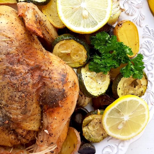 Lemon Pepper Cornish Hen on a white plate with assorted mixed veggies like potatoes, zucchini rounds, mushrooms, and onions on the side. Lemon rounds and parsley for garnish. Whole lemons, garlic cloves, and pepper kernels on the side, white background. Overhead shot. Sugar with Spice Blog.