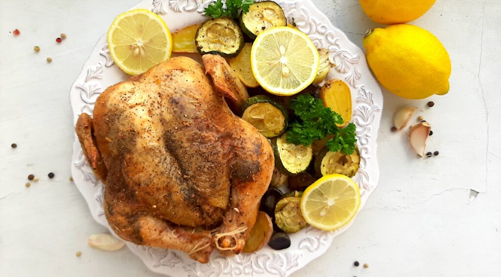 Lemon Pepper Cornish Hen on a white plate with assorted mixed veggies like potatoes, zucchini rounds, mushrooms, and onions on the side. Lemon rounds and parsley for garnish. Whole lemons, garlic cloves, and pepper kernels on the side, white background. Overhead shot. Sugar with Spice Blog.