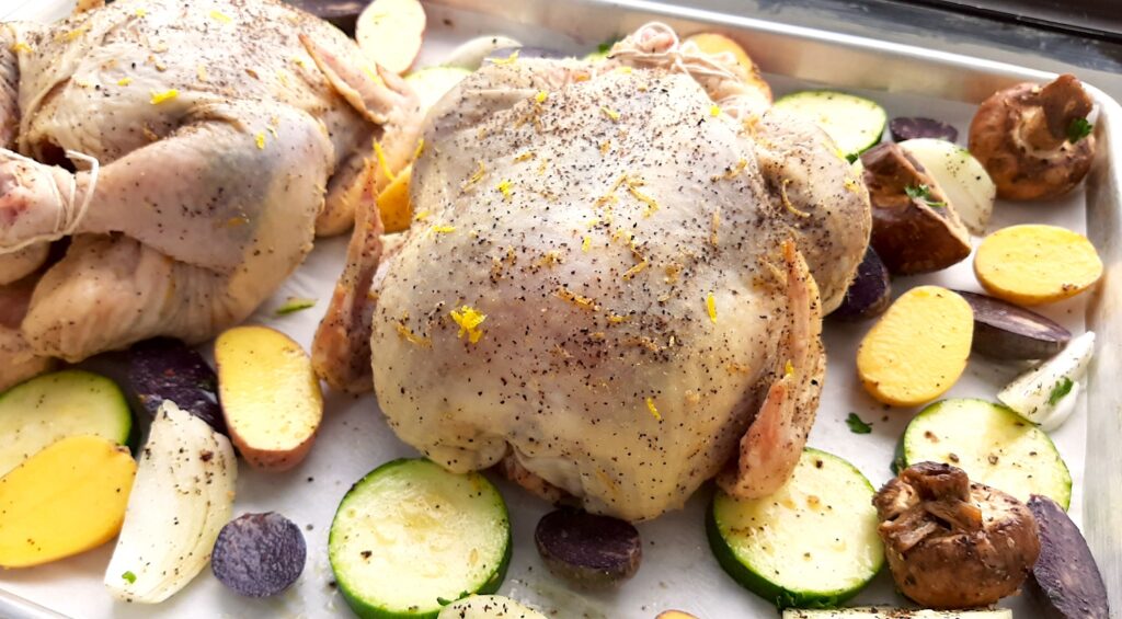 Raw Lemon Pepper Cornish Hen roasted on a sheet pan with assorted mixed veggies like potatoes, zucchini rounds, mushrooms, and onions. Lemon rounds and parsley for garnish. Front shot. Sugar with Spice Blog.