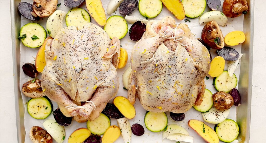 Raw Lemon Pepper Cornish Hen roasted on a sheet pan with assorted mixed veggies like potatoes, zucchini rounds, mushrooms, and onions. Lemon rounds and parsley for garnish. Overhead shot. Sugar with Spice Blog.