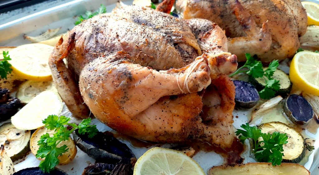 Lemon Pepper Cornish Hen roasted on a sheet pan with assorted mixed veggies like potatoes, zucchini rounds, mushrooms, and onions. Lemon rounds and parsley for garnish. Front shot. Sugar with Spice Blog.