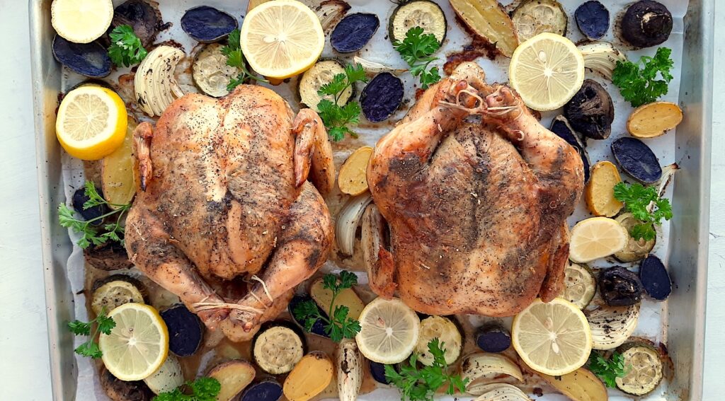 Lemon Pepper Cornish Hen roasted on a sheet pan with assorted mixed veggies like potatoes, zucchini rounds, mushrooms, and onions. Lemon rounds and parsley for garnish. Overhead shot. Sugar with Spice Blog.