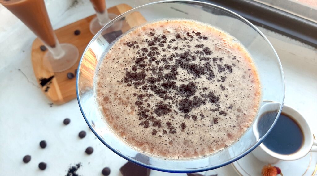 Close up Mocha Martini, Sugar with Spice Blog. Mocha Martini in a large glass on a white background. Two smaller mocha martinis on a wooden cutting board and an espresso in a white cup and saucer in the background with chocolate chips and espresso powder.