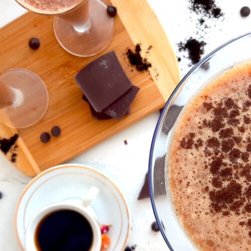 Mocha Martini Recipe Sugar with Spice Blog, One large mocha martini on a white background with espresso powder and chocolate chips in the background. Two smaller mocha martinis on a wooden cutting board with chocolate and an espresso in a white cup and saucer with dried flowers to the side.