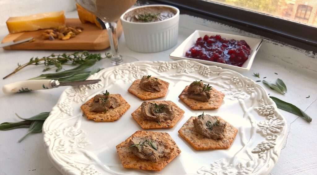 Chicken Liver and Sage Paté smeared on multi seed crackers with thyme leaf garnish on a white decorated plate. Background has more chicken liver and sage paté in a coupe glass and a white ramekin. Cheese board with aged cheeses and pistachios and a dish of lingonberry preserves. Small serving knives, sage leaves, and thyme sprigs scattered around. Front shot. Sugar with Spice Blog.