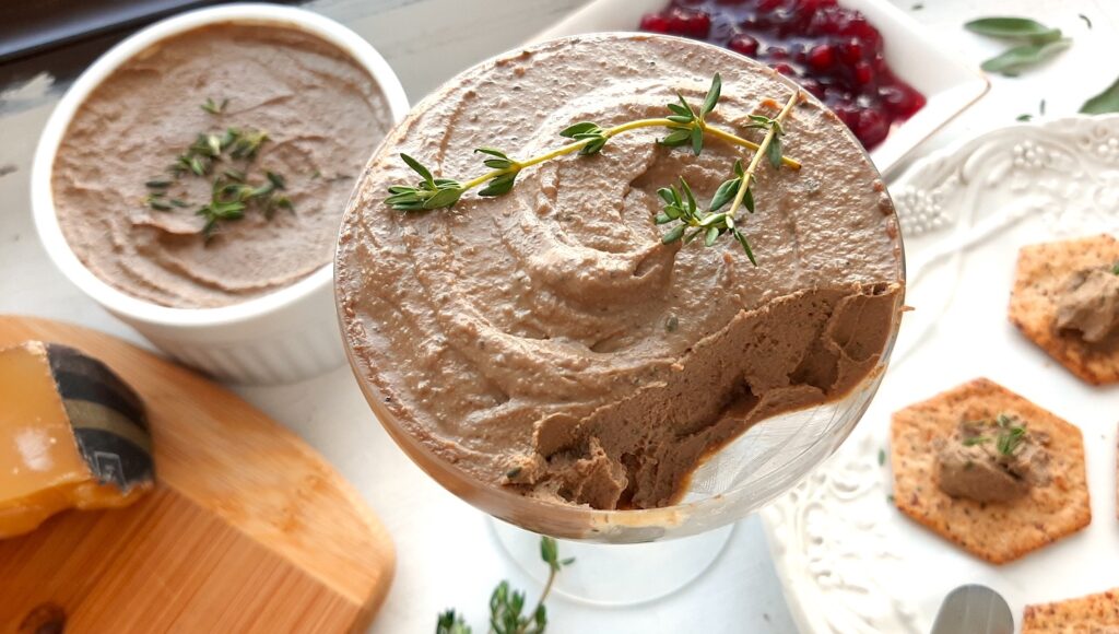 Chicken Liver and Sage Paté in a coupe glass with thyme sprig garnish. Cheese board with aged cheeses and pistachios to one side, white decorated plate with multi-seed crackers smeared with paté to the other side. Background has more chicken liver and sage paté in a white ramekin and a dish of lingonberry preserves. Small serving knives, sage leaves, and thyme sprigs scattered around. Front shot. Sugar with Spice Blog.