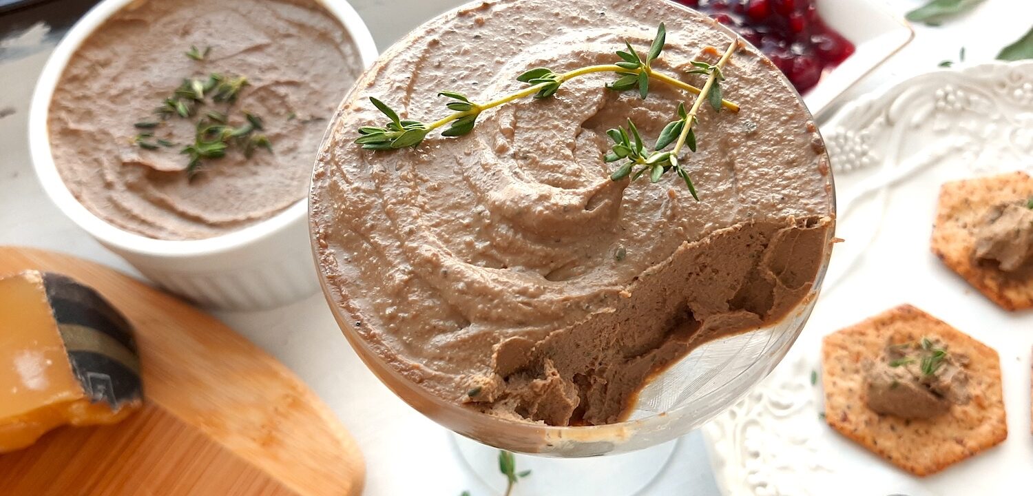 Chicken Liver and Sage Paté in a coupe glass with thyme sprig garnish. Cheese board with aged cheeses and pistachios to one side, white decorated plate with multi-seed crackers smeared with paté to the other side. Background has more chicken liver and sage paté in a white ramekin and a dish of lingonberry preserves. Small serving knives, sage leaves, and thyme sprigs scattered around. Front shot. Sugar with Spice Blog.