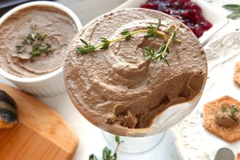 Chicken Liver and Sage Paté in a coupe glass with thyme sprig garnish. Cheese board with aged cheeses and pistachios to one side, white decorated plate with multi-seed crackers smeared with paté to the other side. Background has more chicken liver and sage paté in a white ramekin and a dish of lingonberry preserves. Small serving knives, sage leaves, and thyme sprigs scattered around. Front shot. Sugar with Spice Blog.