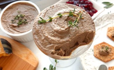 Chicken Liver and Sage Paté in a coupe glass with thyme sprig garnish. Cheese board with aged cheeses and pistachios to one side, white decorated plate with multi-seed crackers smeared with paté to the other side. Background has more chicken liver and sage paté in a white ramekin and a dish of lingonberry preserves. Small serving knives, sage leaves, and thyme sprigs scattered around. Front shot. Sugar with Spice Blog.