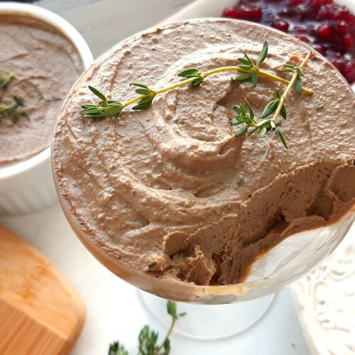 Chicken Liver and Sage Paté in a coupe glass with thyme sprig garnish. Cheese board with aged cheeses and pistachios to one side, white decorated plate with multi-seed crackers smeared with paté to the other side. Background has more chicken liver and sage paté in a white ramekin and a dish of lingonberry preserves. Small serving knives, sage leaves, and thyme sprigs scattered around. Front shot. Sugar with Spice Blog.