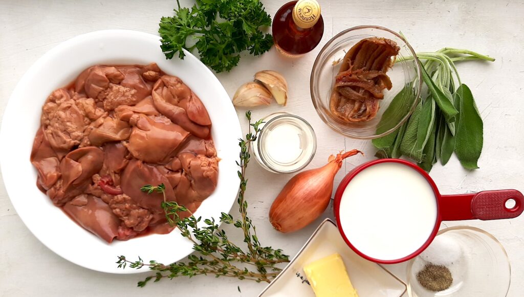 Chicken Liver and Sage Paté ingredients. Raw Chicken Livers in white dish with airplane sized brandy, two garlic cloves, one shallot, a carafe of cream, a pyrex dish of anchovies, a small plate of butter, a cup of milk, fresh parsley, thyme and sage leaves. Overhead shot white background. Sugar with Spice Blog.