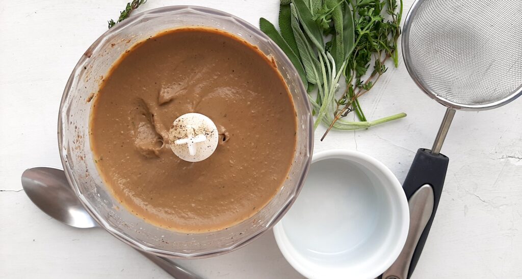 Puréed Chicken Liver and Sage Paté in a blender with a strainer, spoon, ramekin, and fresh herbs surrounding ready to be strained. Overhead shot white background. Sugar with Spice Blog.