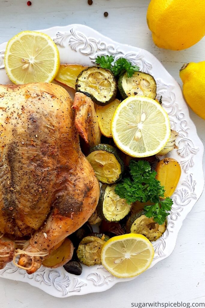 Lemon Pepper Cornish Hen on a white plate with assorted mixed veggies like potatoes, zucchini rounds, mushrooms, and onions on the side. Lemon rounds and parsley for garnish. Whole lemons, garlic cloves, and pepper kernels on the side, white background. Overhead shot. Pinterest Image. Sugar with Spice Blog.