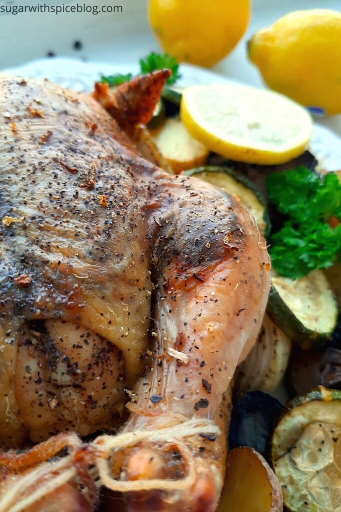 Lemon Pepper Cornish Hen on a white plate with assorted mixed veggies like potatoes, zucchini rounds, mushrooms, and onions on the side. Lemon rounds and parsley for garnish. Whole lemons, garlic cloves, and pepper kernels on the side, white background. Front shot. Pinterest Image. Sugar with Spice Blog.