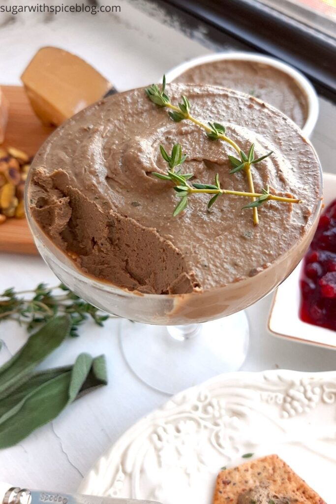 Chicken Liver and Sage Paté in a coupe glass with thyme sprig garnish. Cheese board with aged cheeses and pistachios to one side, white decorated plate with multi-seed crackers smeared with paté to the other side. Background has more chicken liver and sage paté in a white ramekin and a dish of lingonberry preserves. Small serving knives, sage leaves, and thyme sprigs scattered around. Front shot. Sugar with Spice Blog.