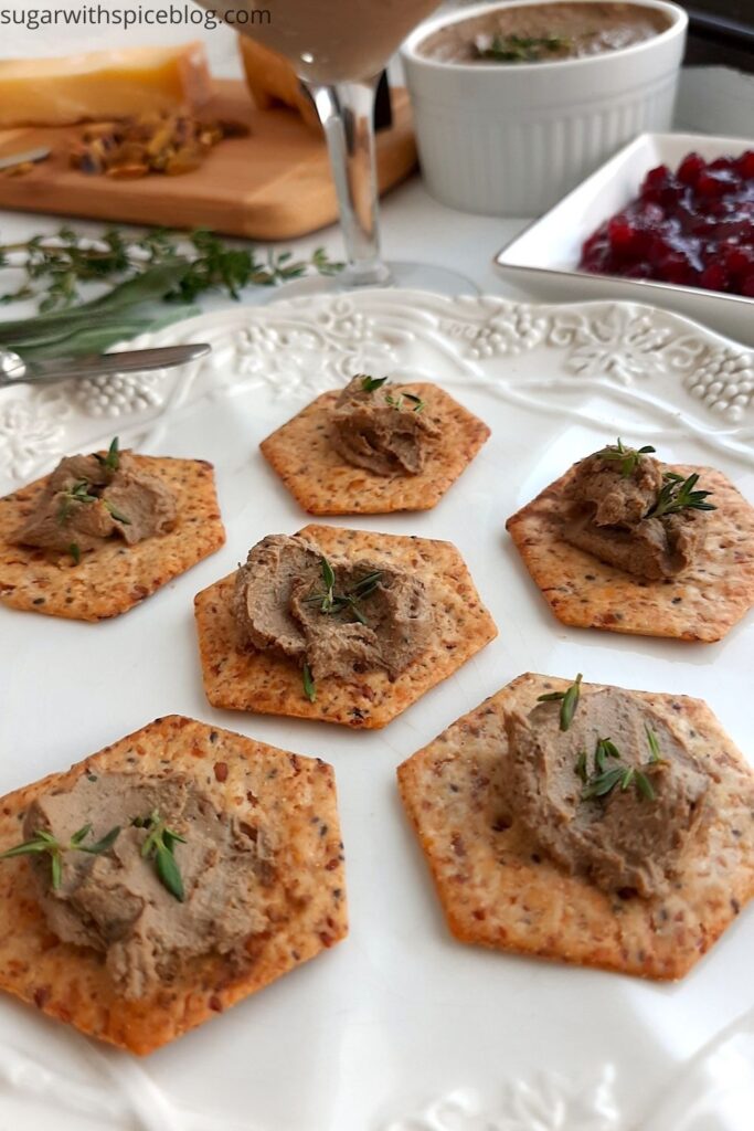 Chicken Liver and Sage Paté smeared on multi seed crackers with thyme leaf garnish on a white decorated plate. Background has more chicken liver and sage paté in a coupe glass and a white ramekin. Cheese board with aged cheeses and pistachios and a dish of lingonberry preserves. Small serving knives, sage leaves, and thyme sprigs scattered around. Front shot. Pinterest Image. Sugar with Spice Blog.