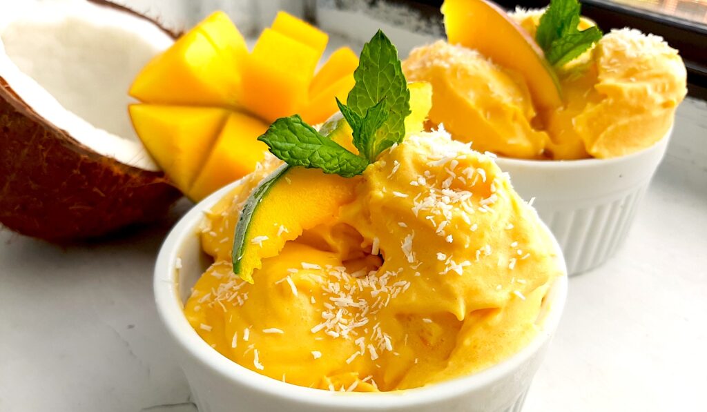 Homemade mango coconut soft serve ice cream in a white ramekin topped with shredded coconut and mint sprigs. Surrounded by halved coconut, sliced mangos and a second cup of ice cream. On a white background front shot. Sugar with Spice Blog.