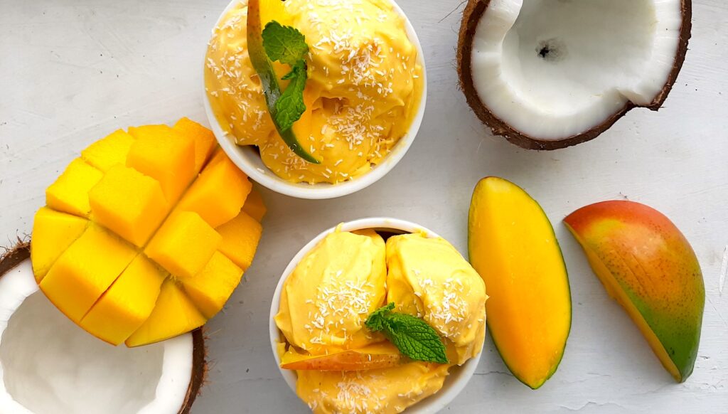 Two homemade mango coconut soft serve ice creams in white ramekins topped with shredded coconut and mint sprigs. Surrounded by halved coconut and sliced mangos. On a white background overhead shot. Sugar with Spice Blog.